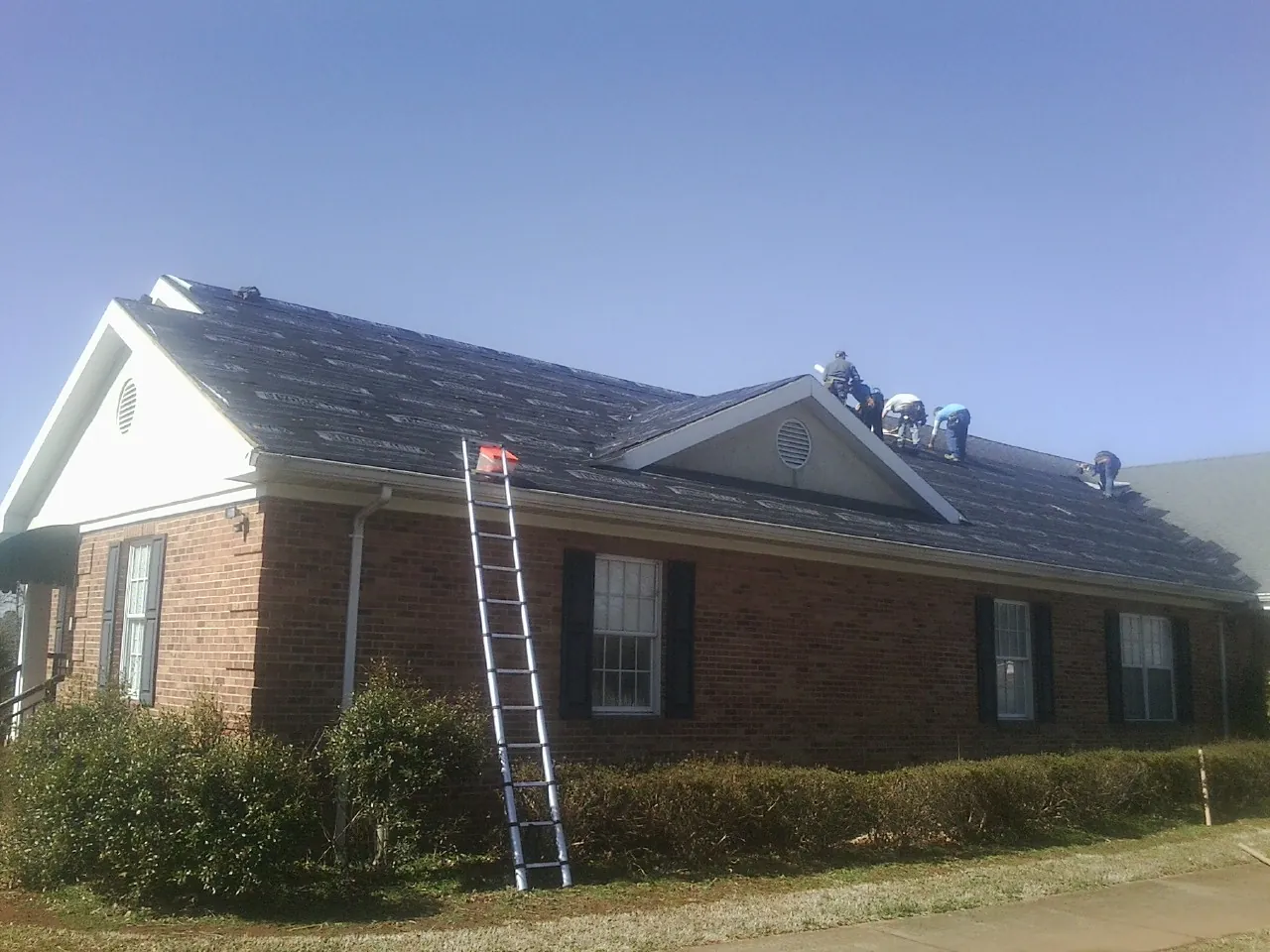 Crew tearing shingles off a brick ranch roof with a ladder set — Emergency Roof Repair work by our crew in Mc Intosh