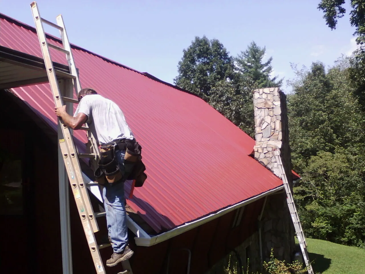 Roofer on a ladder beside a red metal roof and stone chimney — Soffit & Fascia Repair by Roofing Brothers in Mc Intosh
